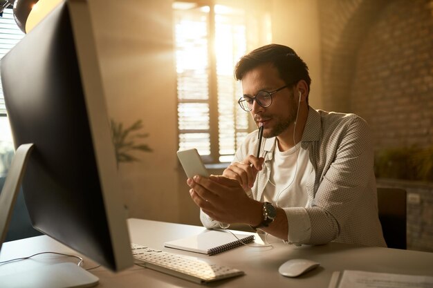 彼のオフィスの机でコンピューターで作業している間、携帯電話で物思いにふける起業家のテキストメッセージ