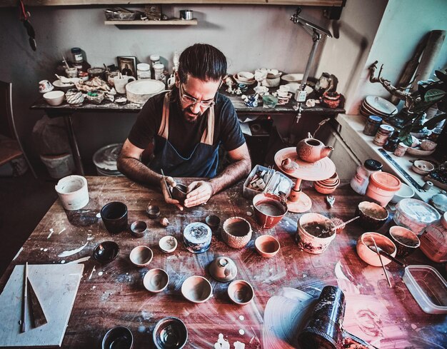 Задумчивый гончар-энтузиаст в очках в собственной мастерской работает над новым чайником ручной работы.