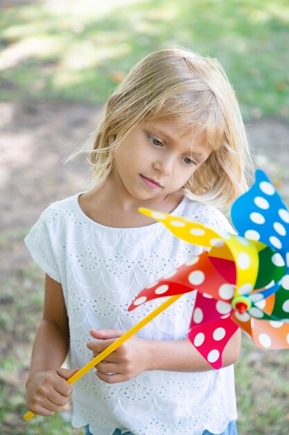 Pensive fair haired girl standing in park, holding pinwheel, looking at toy. Vertical shot. Children outdoor activity concept