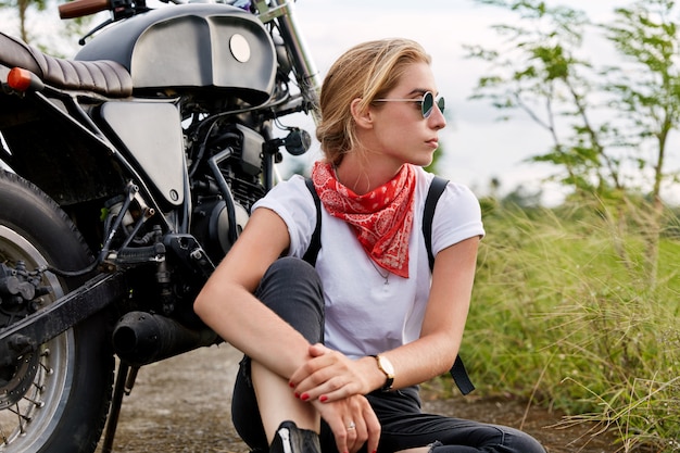 Pensive female biker deep in thoughts wears fashionable clothing, looks away with thoughtful expression, sits on ground near motorbike, covers long destination. People, transport and freedom
