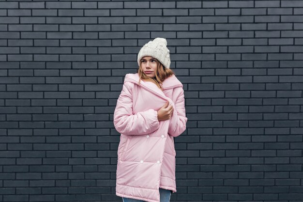Pensive female model in winter clothes posing in cold day. Enchanting caucasian woman in hat standing on gray brick wall.