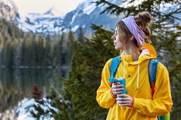 物思いにふける女性登山家は頭の周りにスカーフ、黄色のレインコートを着て、フラスコからのホットコーヒーを楽しんでいます