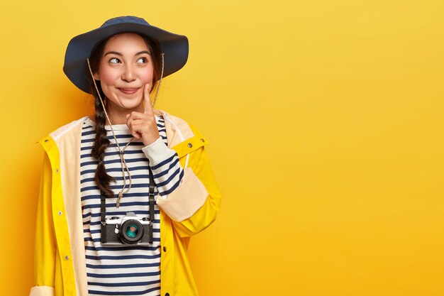 Pensive female tourist keeps index finger on cheek, thinks what way to choose, explores local surrounding during hiking trip, wears retro camera on neck, headgear and yellow waterproof raincoat