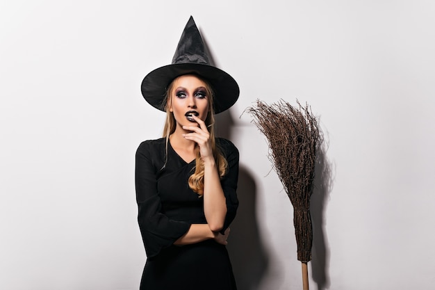 Задумчивая женщина-волшебник позирует на белой стене. Чувственная молодая ведьма в черной шляпе, стоя рядом с метлой.