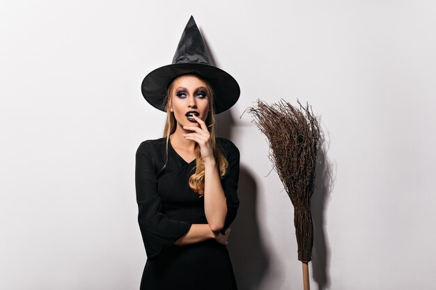 Pensive female wizard posing on white wall. Sensual young witch in black hat standing beside broom.