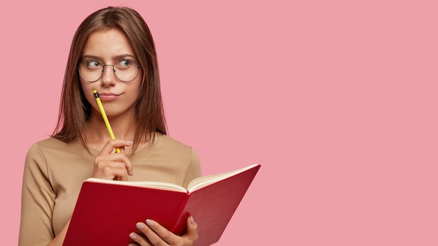 Free photo pensive female writer ponders on new ideas, holds red notebook and pencil near mouth, has thoughtful expression