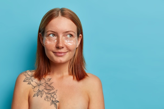 Free photo pensive freckled woman with natural red hair, stands shirtless, looks aside and thinks about something pleasant, wears transparent patches for reducing puffiness, isolated on blue wall