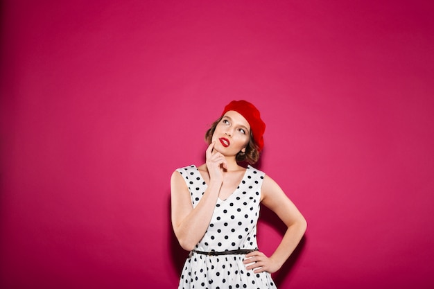 Pensive ginger woman in dress with arm on hip holding her chin and looking up over pink