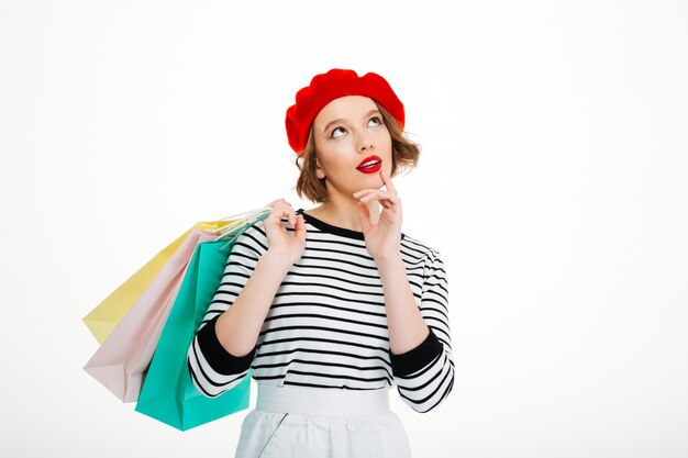 Pensive ginger woman holding packages and looking up while holding her chin over grey