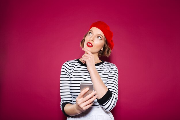 Pensive ginger woman holding smartphone and looking up over pink