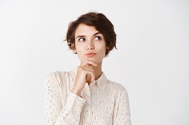 Pensive girl in blouse touching chin, looking at upper right corner and thinking, making choice, standing over white wall