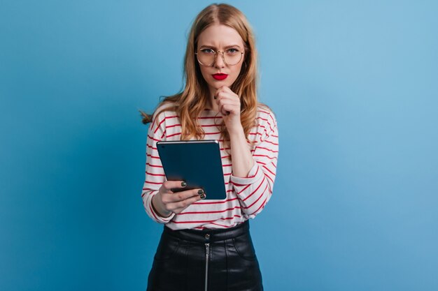 デジタルタブレットを保持している眼鏡の物思いにふける女の子。青い背景にガジェットを使用して金髪の女性のスタジオショット。