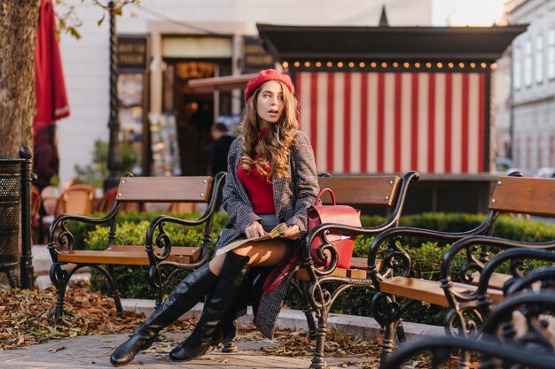 Pensive girl in knee high boots sitting on bench and looking away with mouth open