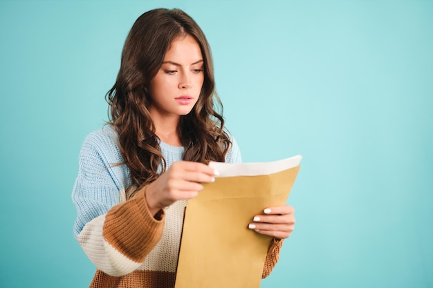 Free photo pensive girl in knitted sweater thoughtfully opening response envelope over blue background