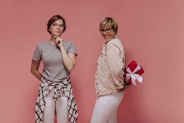 Pensive girl in plaid shirt and glasses posing with smiling woman in white and beige clothes with gift box on isolated background.