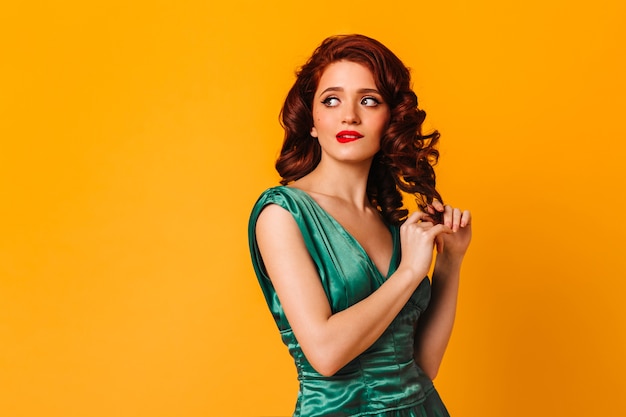 Pensive girl touching curly hair on yellow space. Studio shot of thoughtful woman in green dress.