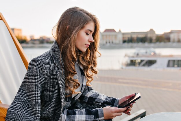 Pensive girl with curly hairstyle texting message, sitting on recliner in cold morning