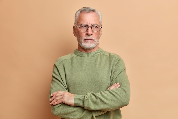 Pensive grey haired bearded old man stands with arms crossed and looks away thoughtfully wears casual jumper ponders on plans for weekend going to visit children isolated over brown studio wall