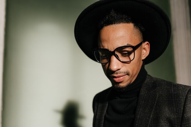 Pensive guy in glasses looking down. Indoor photo of sad african man in hat.