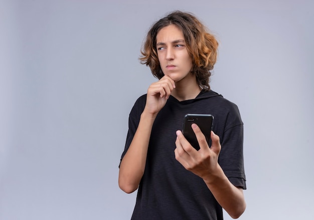 Pensive guy with long hair in black t-shirt holding a phone on white wall