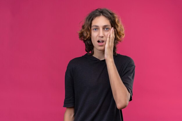 Pensive guy with long hair in black t-shirt put his hand on cheek on pink wall