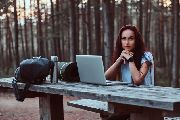 Pensive hipster girl in white shirt looking at camera while sitting on a wooden bench with an open laptop in a beautiful autumn forest.