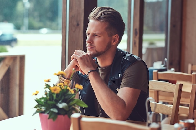 Hipster pensieroso con un taglio di capelli alla moda e la barba si siede a un tavolo in un caffè lungo la strada, guarda la strada.