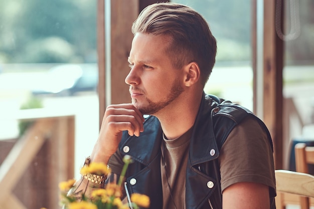 Pensive hipster with a stylish haircut and beard sits at a table in a roadside cafe, looks at the street.