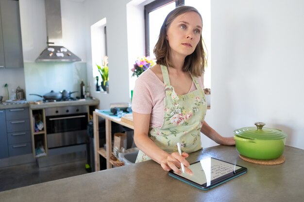 物思いにふける主婦が毎週のメニューを計画し、彼女のキッチンで食料品のリストを書き、カウンターの大きな鍋の近くにあるタブレットを使用して、見上げる。家庭料理とオンライン料理のコンセプト