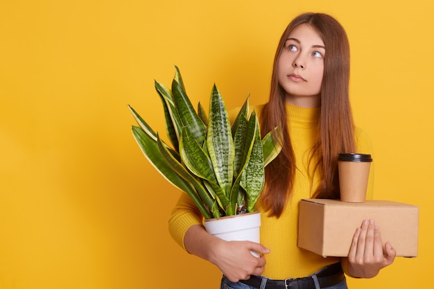 Pensive lady dresses casual clothing holding her staff and flowerpot in hands, looking aside, thinks about her dismissal, being sad, looks thoughtful, standing isolated over yellow wall.