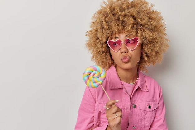 Free photo pensive lovely woman with curly bushy hair keeps lips folded wears trendy heart shaped sunglasses holds sweet caramel candy dressed in pink jacket isolated over white background empty space