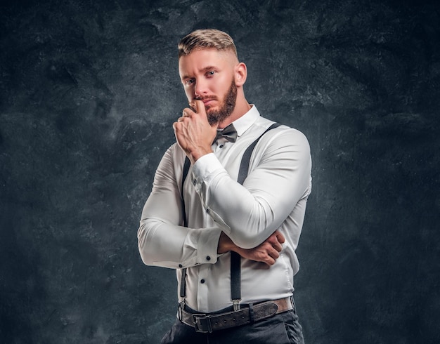 Maschio pensieroso che pensa a qualcosa di importante. giovane elegantemente vestito in camicia con farfallino e bretelle in posa con la mano sul mento. foto in studio su sfondo scuro della parete