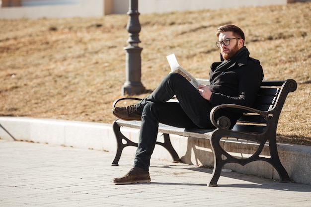 Задумчивый мужчина сидит на скамейке и читает газету на открытом воздухе