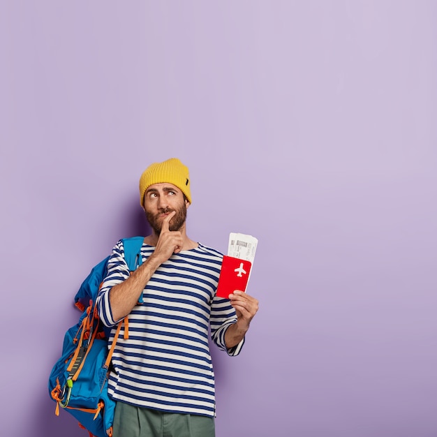 Free photo pensive man traveler holds chin, focused upwards, plans future trip abroad, holds passport with flying boarding pass, carries rucksack