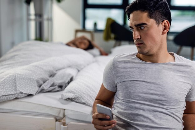 Pensive man using mobile phone and cheating on his girlfriend who is sleeping on the bed behind him