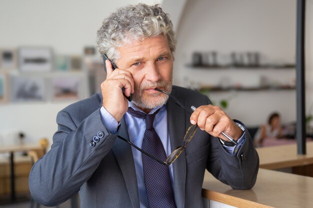 Pensive mature businessman talking on mobile phone, standing at co-working, leaning on desk, looking at camera a