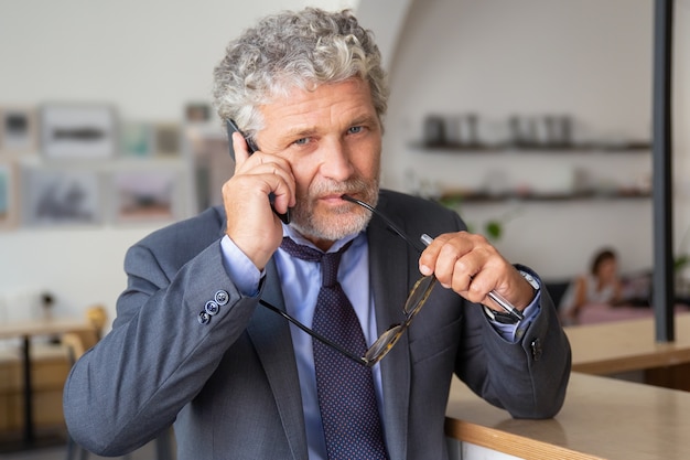 Pensive mature businessman talking on mobile phone, standing at co-working, leaning on desk, looking at camera a