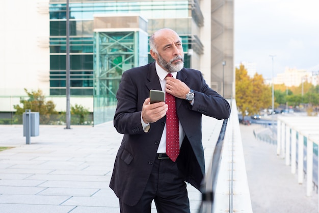 Pensive mature executive adjusting tie and using cellphone