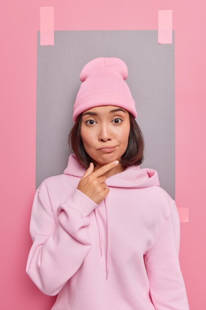 수심에 찬 밀레니엄 아시아 여성은 캐주얼한 후드티를 입은 흥미로운 제안과 회반죽이 있는 종이로 분홍색 벽에 포즈를 취하는 흥미로운 제안을 고려하는 의심스러운 표정을 하고 있습니다.
