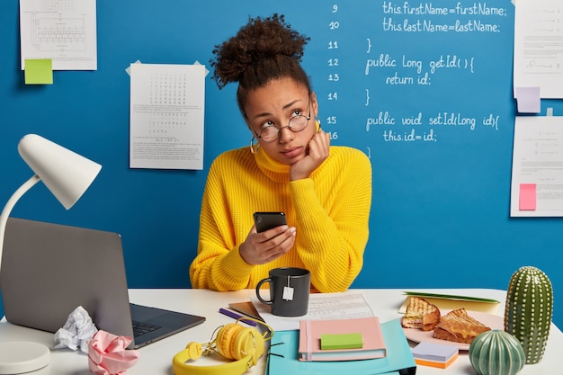 Free photo pensive office worker thinks on creative idea, uses mobile phone, waits for call, prepares for course work, learns languages online, spends time for autodidact drinks tea wears yellow sweater