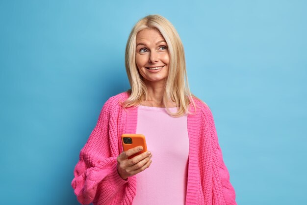 Pensive pleased cheerful blonde woman with pleasant appearance uses mobile phone for online communication dressed in warm knitted pink jumper.