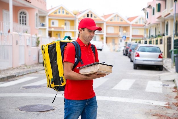 Pensive postman holding parcel and reading address in order sheet. attractive deliveryman in red cap and shirt standing outdoors