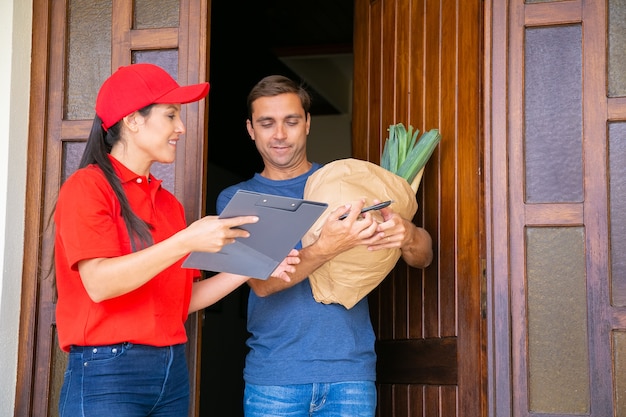 Pensive postwoman holding clipboard and showing data in order sheet. Attractive customer standing, receiving vegetables in paper bag from grocery store. Food delivery service and post concept