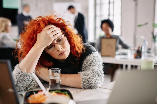 Free photo pensive redhead entrepreneur feeling tired while sitting at her desk in the office there are people in the background