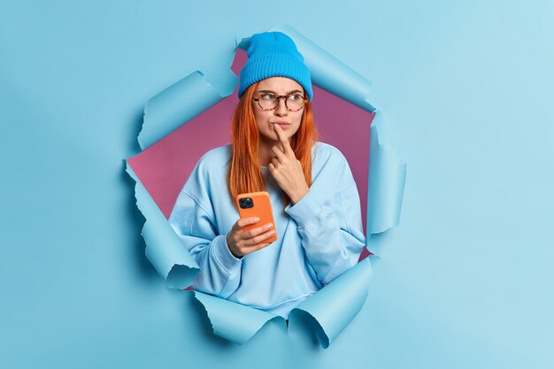 Pensive redhead woman holds mobile phone with thoughtful expression keeps index finger near lips browses in internet thinks over received message wears hat and jumper breaks through blue paper wall