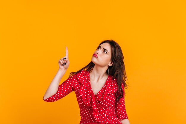 Pensive sad brunette in red top shows finger up. Portrait of girl on isolated yellow wall.