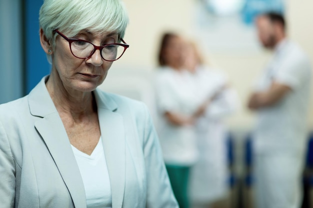 Free photo pensive senior woman standing in a lobby in the hospital and thinking of something group of doctors are in the background