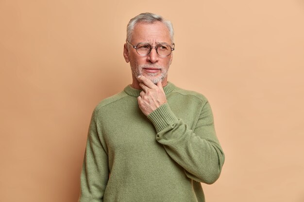 Pensive serious old bearded man holds chin and looks away thoughtfully thinks over received suggestion contemplates about something considers wears casual jumper isolated over brown wall