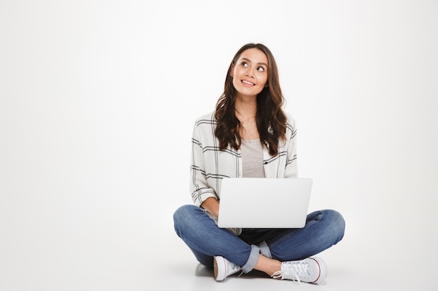 Pensive smiling brunette woman in shirt sitting on the floor with laptop computer and looking up over gray