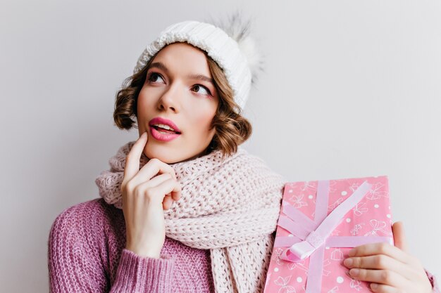Pensive spectacular girl in cute hat posing with pink present box.  ecstatic woman wears knitted scarf thinking about something, holding new year gift.